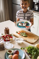 Laughing male kid cooking breakfast decorating fresh sandwich with berries and fruits at kitchen