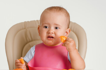 A baby in a pink bib sits on a chair and eats tangerines, a grimace of disgust at the sour taste of citrus fruits