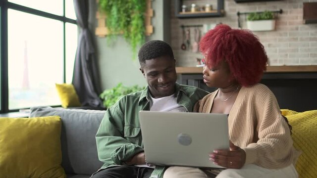 Young couple in love using laptop and hugging while sitting on couch in home room spbd. African American man, woman look at computer screen with smiles and talk, search Internet and sit on sofa in