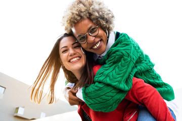 Playful multiracial female lesbian couple looking at camera. Black woman piggyback ride with girlfriend. Copy space.
