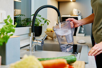 Woman pouring water from faucet into water filter jug at the kitchen - obrazy, fototapety, plakaty