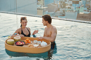 Smiling pretty young couple enjoying breakfast in rooftop swimming pool