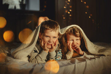 Christmas photo. Children, brother and sister at home on the bed under a warm blanket are smiling happily, laughing. Around Christmas lights, garlands, bokeh.