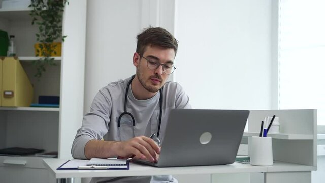 Doctor at distance appointment with internet resources. Skilled male internist in spbd uniform writes notes and answers videocall via modern laptop in clinic