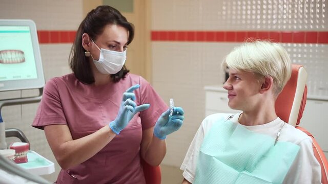 Teenager visits orthodontist. Brunette doctor in mask and gloves shows spbd removable aligner model to young blond patient at appointment in clinic office
