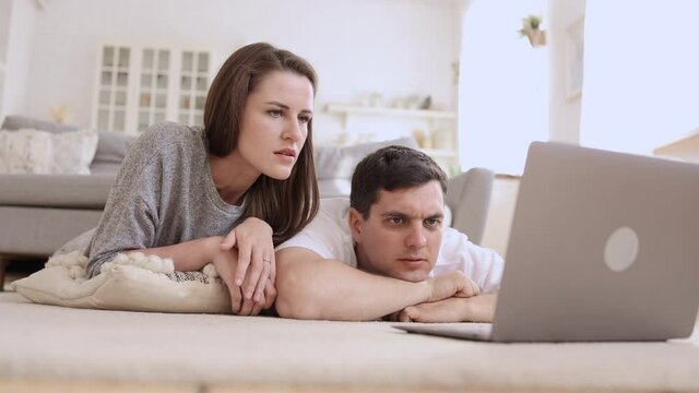 Young couple watching web or video and using device while lying on floor in apartment room spbi. American woman, man look at computer screen and are surprised, talk and lie in light interior. 4k video