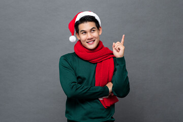 Smiling handsome Asian man wearing  Christmas theme clothes looking at empty space aside and thinking with hand pointing up, studio shot isolated on gray background