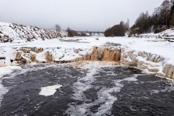 Winter view of the river valley and waterfall. Freezing river. The ice and the banks are covered with snow. Cold winter weather. Famous tourist attraction of the Leningrad region. Tosna river, Russia.