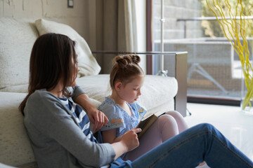 Obraz premium Smiling young mother and little daughter sitting on floor near sofa at home and reading books.