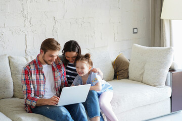 Positive friendly young parents with smiling little daughter sitting on sofa together watching photos on laptop, relaxing at home on weekend.