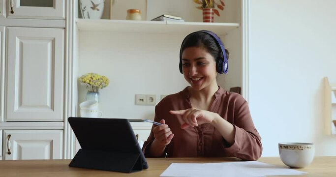 Energetic young Indian female work from home office sit by digital tablet in wireless headset start remote conversation online. Positive millennial woman teacher webcast live record educational vlog
