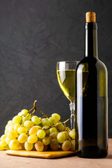 a glass glass and a bottle of white wine and a brush of white grapes on a cutting board on a black background