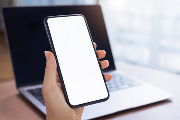 Close up of hand holding smartphone with white mock-up screen and laptop..