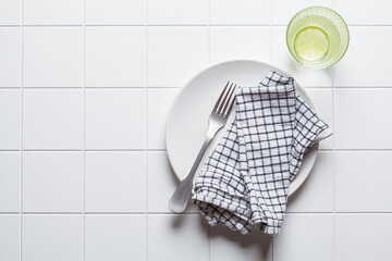 Empty white plate with fork, napkin and glass of water on white tile background, copy space, top view. Intermittent fasting, healthy lifestyle, diet concept.