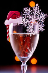 a glass of champagne with a decoration in the form of a Santa Claus hat, a caramel cane and snowflakes on a black background with a backlit close-up