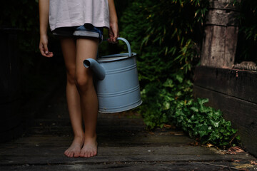 Low section of little girl in shorts holding watering can outdoors in garden.