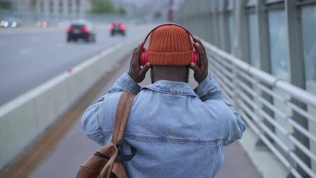 Contemporary gadgets to enjoy audio. African-American man tourist puts on bluetooth headphones spbas walking along road pavement past building backside view