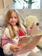 A girl in an embroidered shirt with a book in her hands. A large teddy bear is sitting nearby.