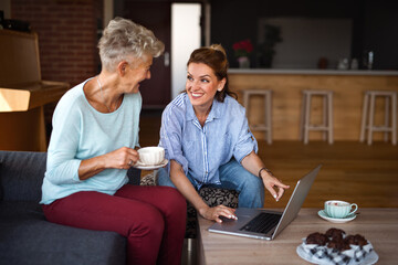 Senior mother having coffee with adult daughter indoors at home, sitting, talking and using tablet.