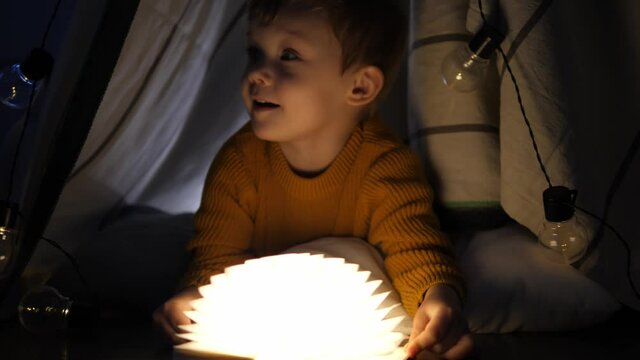 Little happy boy playing with a book sitting on a cozy tent.