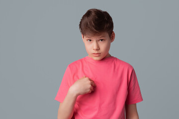 Indignant displeased teenager boy points at herself asks why me, dressed in casual pink t shirt, poses against gray background indicates self