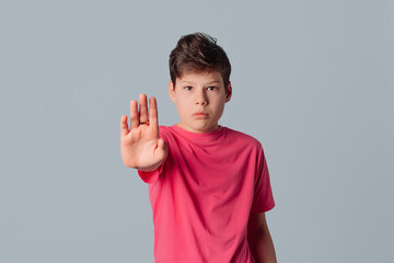 Serious teenager boy in casual pink t shirt raise hand in block, say no, standing against gray...