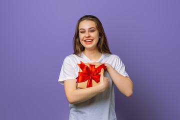 Fototapeta premium Picture of happy young brunette woman in casual white t shirt with gift box and looking at the camera, standing isolated over purple background. New Year Women's Day birthday holiday concept