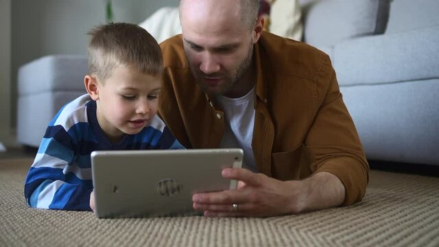 Father and toddler son have fun in internet. Careful baldhead man with spbd lovely little boy use modern tablet lying on floor carpeting near couch closeup