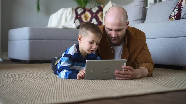 Father and toddler online development education. Lovely little boy child spbd does tasks on modern tablet while parent lies nearby on floor in living room