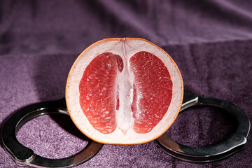 Half grapefruit and handcuffs close-up on a purple background. The concept of protecting virginity, sexual inviolability. Vagina symbol. - obrazy, fototapety, plakaty