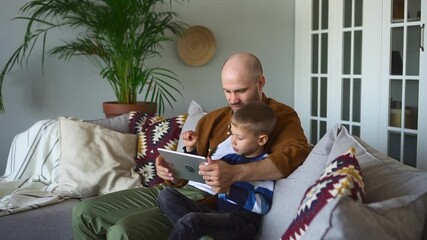 Father and toddler son have fun at home. Loving bald parent hugs spbd little child doing developing tasks on tablet sitting together on couch in living room