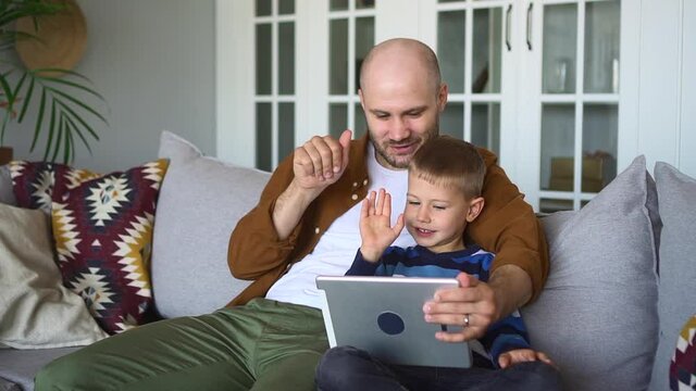 Father and son at videochat. Positive parent and funny little child send air kisses to mother finishing call via tablet sitting on couch in living room spbd