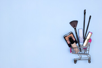 Various make-up products and brushes in shopping cart on blue background. Makeup cosmetics sale...