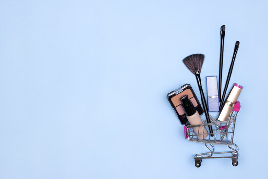 Various make-up products and brushes in shopping cart on blue background. Makeup cosmetics sale concept. Copy space