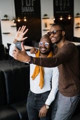 Two African friends of the black race, fashionably dressed, standing against the background of a cafe with a dark interior, take selfies and communicate online via a smartphone. The Concept of Friends