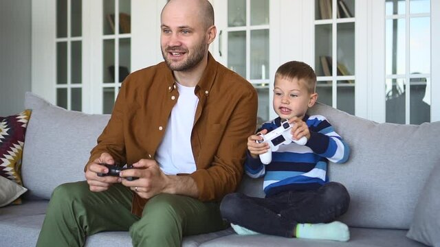 Father spends time with little son. Joyful baldhead man and funny blond child with joysticks play videogame sitting on grey sofa against bookcase at home spbd