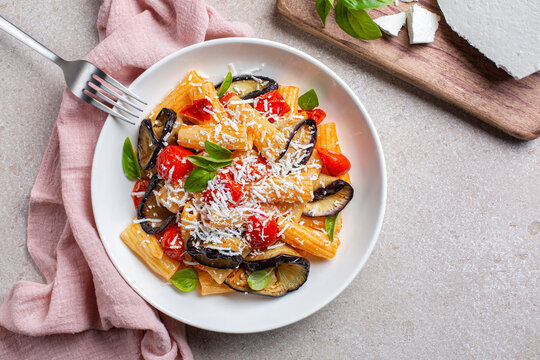 Italian vegetarian pasta with eggplant, tomato sauce, basil, served with grated ricotta salata cheese in a white plate. Pasta salad. Pink and beige table surface. Directly above.