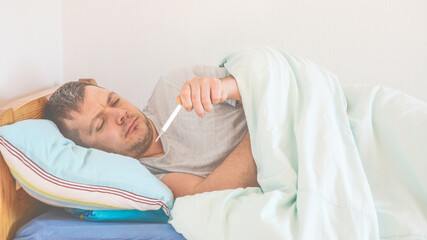 Fototapeta na wymiar Sick man with a Thermometer in the Bed at the Home. Concept of illness, fever symptoms