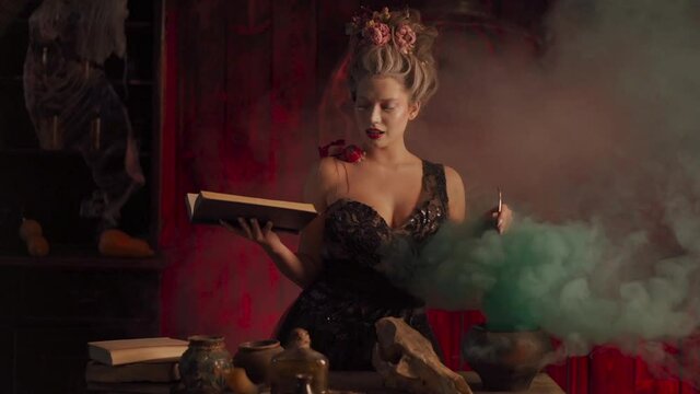 Halloween witch with cauldron and magic book. Beautiful woman flowers in hair conjuring, making witchcraft. Standing spooky dark room. Enchantress prepare love potion use magic spell. Handheld