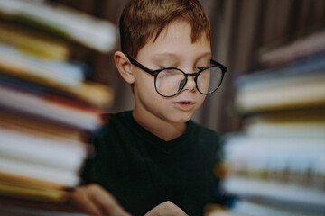cute caucasian boy wearing glasses covering head with book. Cheerful boy peeking from behind piles...