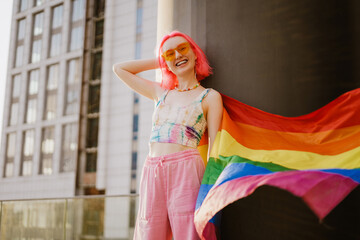 Young white woman laughing while standing with rainbow flag by wall