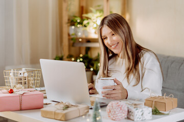 Happy and smiling young woman wrapping and preparing Christmas gifts, typing at laptop and having online video chat. Online shopping at Christmas holidays. Home interior with lights and plants
