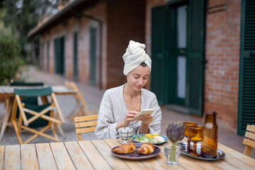 Woman using smartphone and having breakfast during resting in hotel at morning time. Concept of weekend, tourism and vacation. Young caucasian woman wearing bathrobe and wrapped bath towel on head