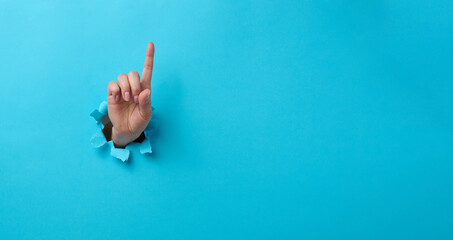 female hand sticking out of a torn hole in a blue paper background, attention gesture. Place for an...