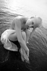 Art nude sexy blonde with short haircut is sitting in water on shore beach of lake at sunset. Wet hair and a woman body. Secluded beach holiday. Black and white