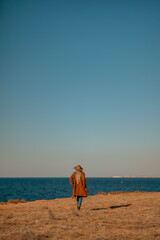 A woman walking along the coast near the sea. An elegant lady in a brown coat and a hat with fashionable makeup walks on the seashore