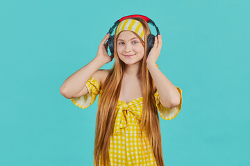 Studio portait of Funny young blonde woman in yellow dress isolated on blue background. Girl Listen music with wireless headphones using music streaming service.