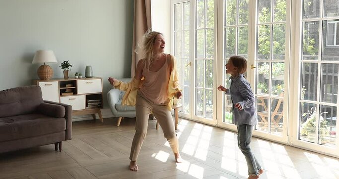 Two cute energetic sisters grownup older and younger schoolgirl enjoy dancing at living room on floor with underheating by picture window. Happy young mom little daughter celebrate moving to new flat