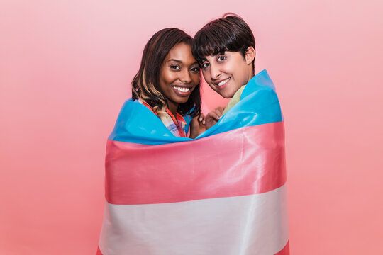 Smiling girlfriends covered in transgender flag by pink background