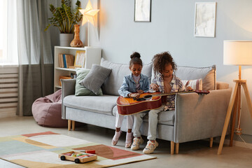 Full length portrait of two cute girls playing with acoustic guitar in cozy home interior, copy...